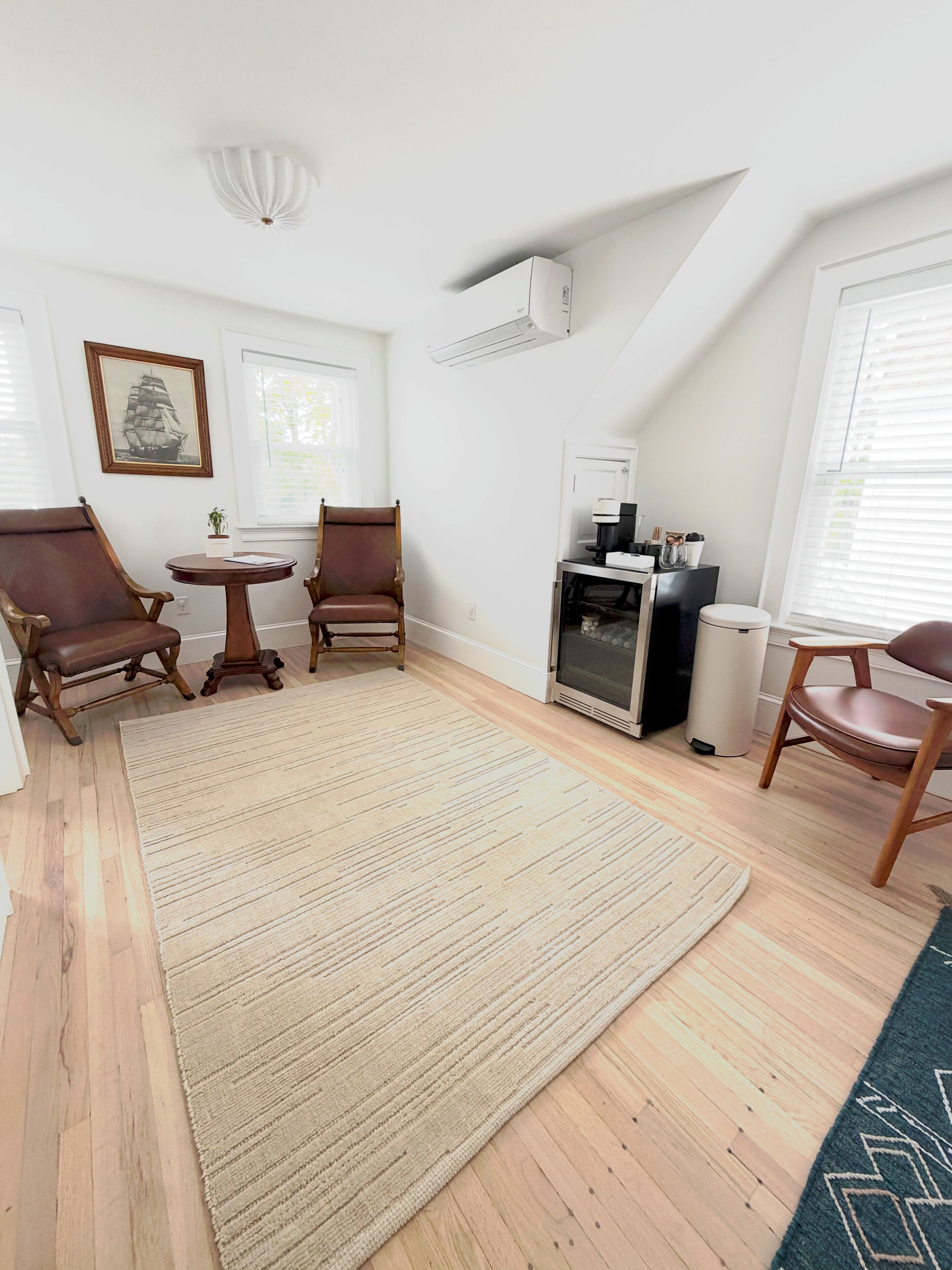 A room with a large rug, two wooden chairs, a small table, a fireplace, and a framed picture on the wall.