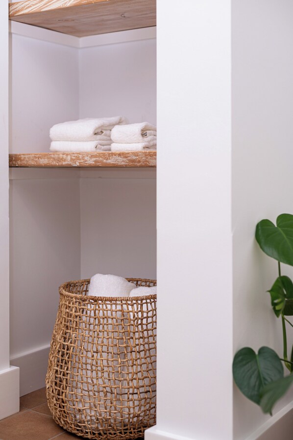 A corner of a room with a wooden shelf holding folded towels, a wicker basket with towels, and a potted plant.