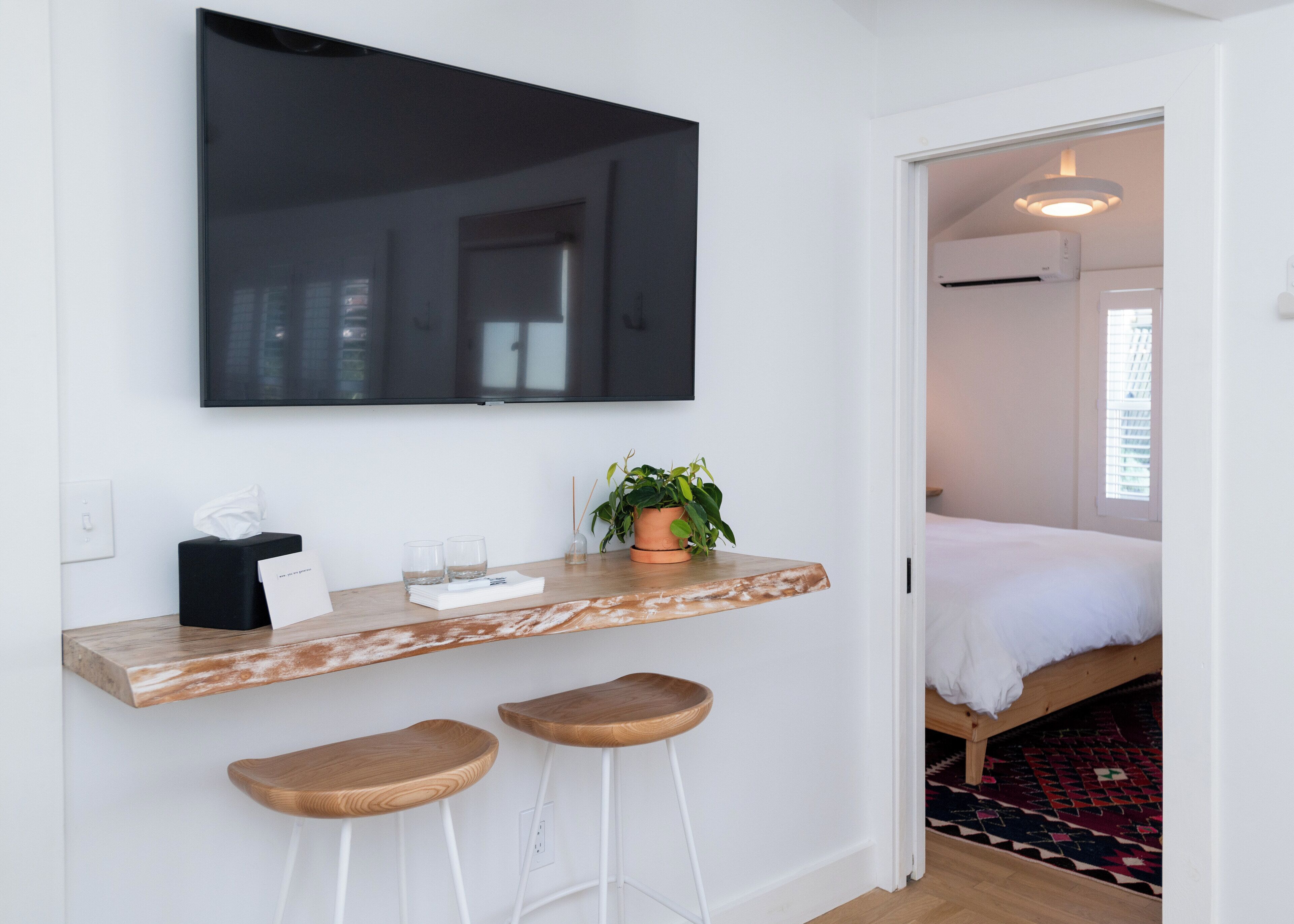 Una habitación moderna con una televisión de pantalla plana grande montada en la pared, una barra de madera con dos taburetes, una planta en maceta y una cama con un tapete estampado.