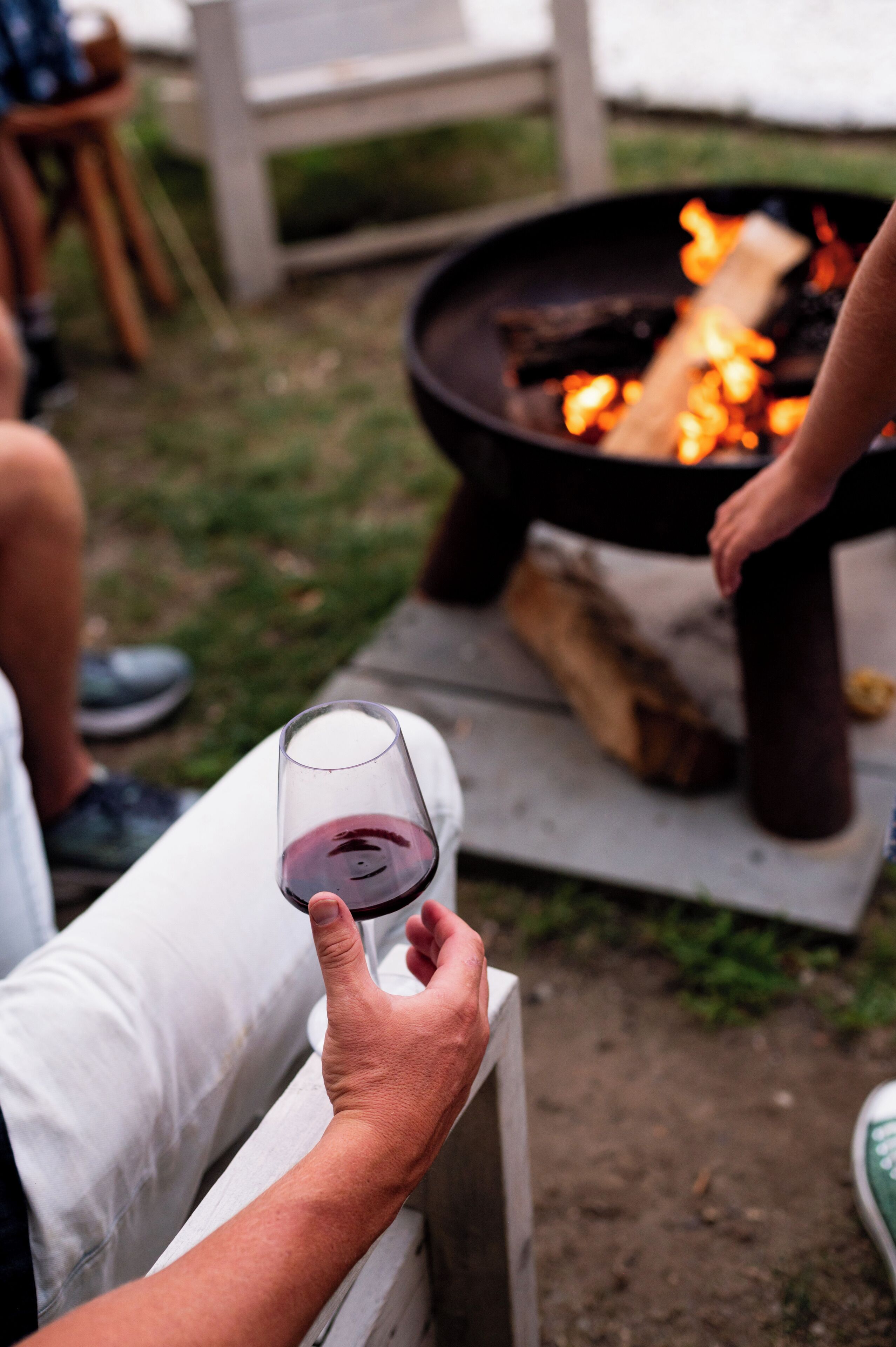 A person holding a glass of red wine, sitting near a fire pit with logs and flames.