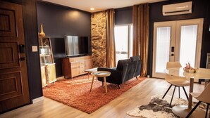 A modern living room with a flat-screen TV, a black sofa, a dining table with chairs, and a stone accent wall. - Suite 101 is the Main House's largest and most luxurious suite. (Cherokee Village)