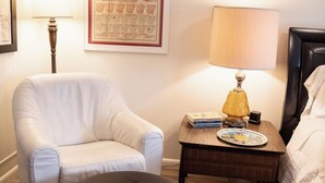 A room with a white armchair, a black ottoman, a wooden nightstand with a lamp, a framed map of Arkansas, and a bed with white bedding. - Suite 103 also known as the Arkansas Suite (Cherokee Village)