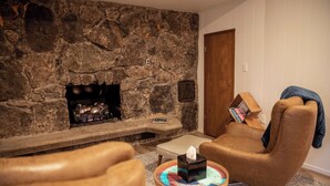 A stone fireplace, a leather armchair, a wooden side table with a tissue box, and a bookshelf. - Suite 105 freatures a private outdoor-covered balcony. (Cherokee Village)