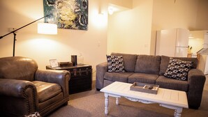 A living room with a leather armchair, a grey sofa with patterned cushions, a white coffee table, and a painting on the wall.