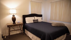 A neatly made bed with a dark blue bedspread, a pillow with 'welcome' text, a bedside lamp, and a small table with a clock.