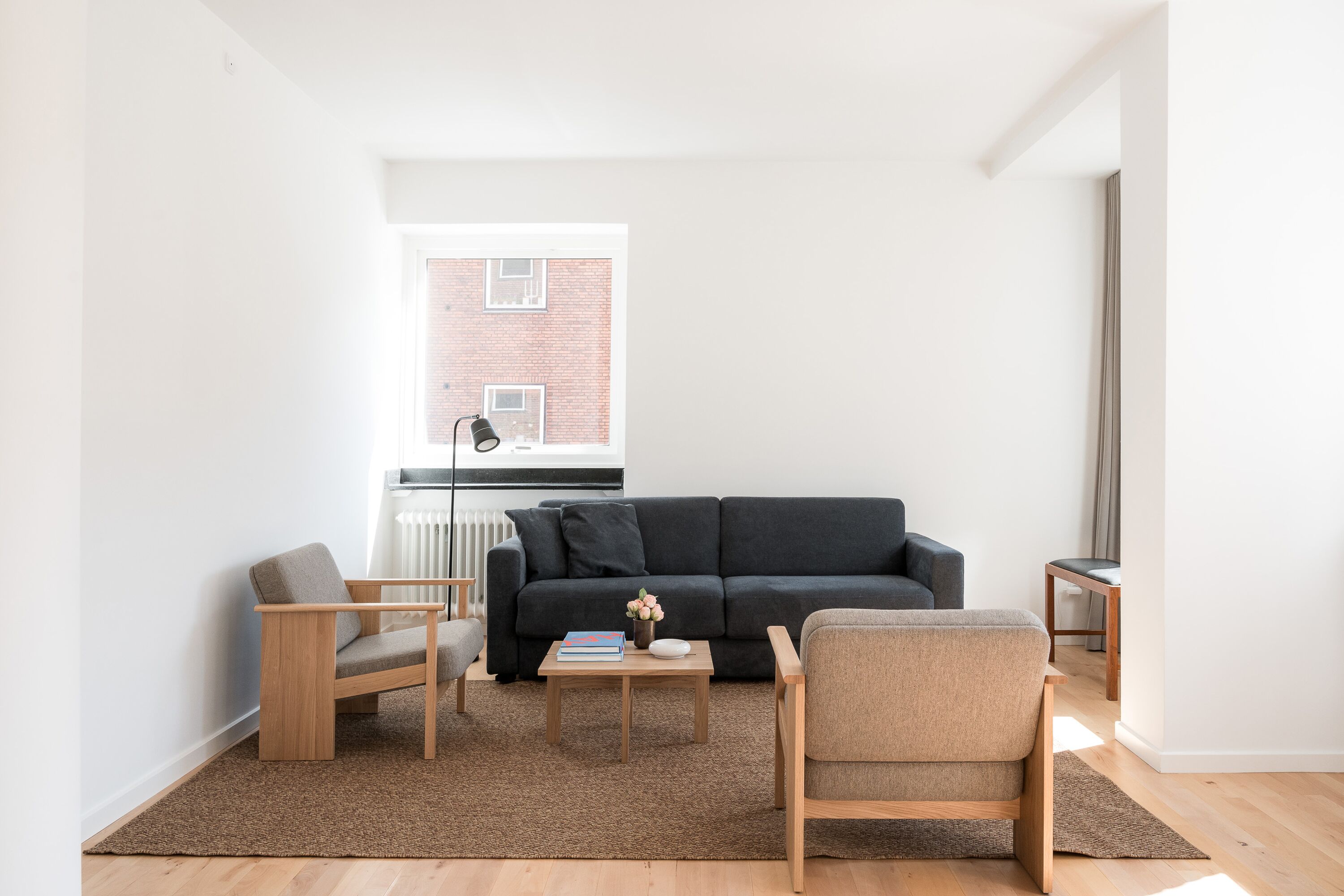 Ein modernes Wohnzimmer mit einem schwarzen Sofa, zwei Holzstühlen, einem Couchtisch mit Büchern und Blick auf ein Ziegelgebäude durch ein Fenster.