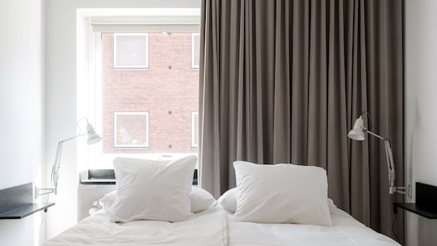 A neatly made bed with white bedding, a window with blinds, and a curtain. - Queens Courtyard (Copenhagen)