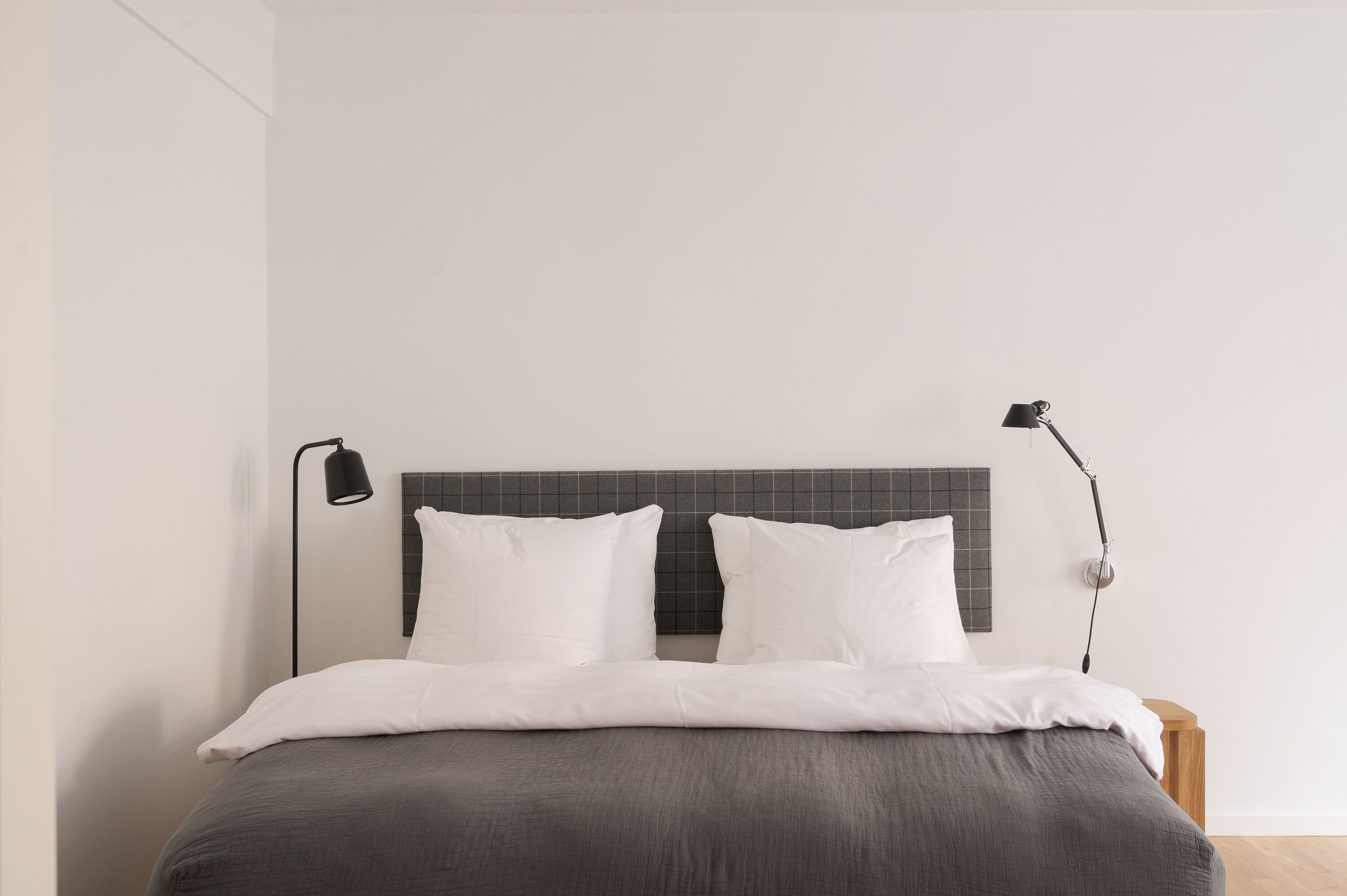 A neatly made bed with a grey bedspread and white pillows, a black floor lamp, and a wooden nightstand.
