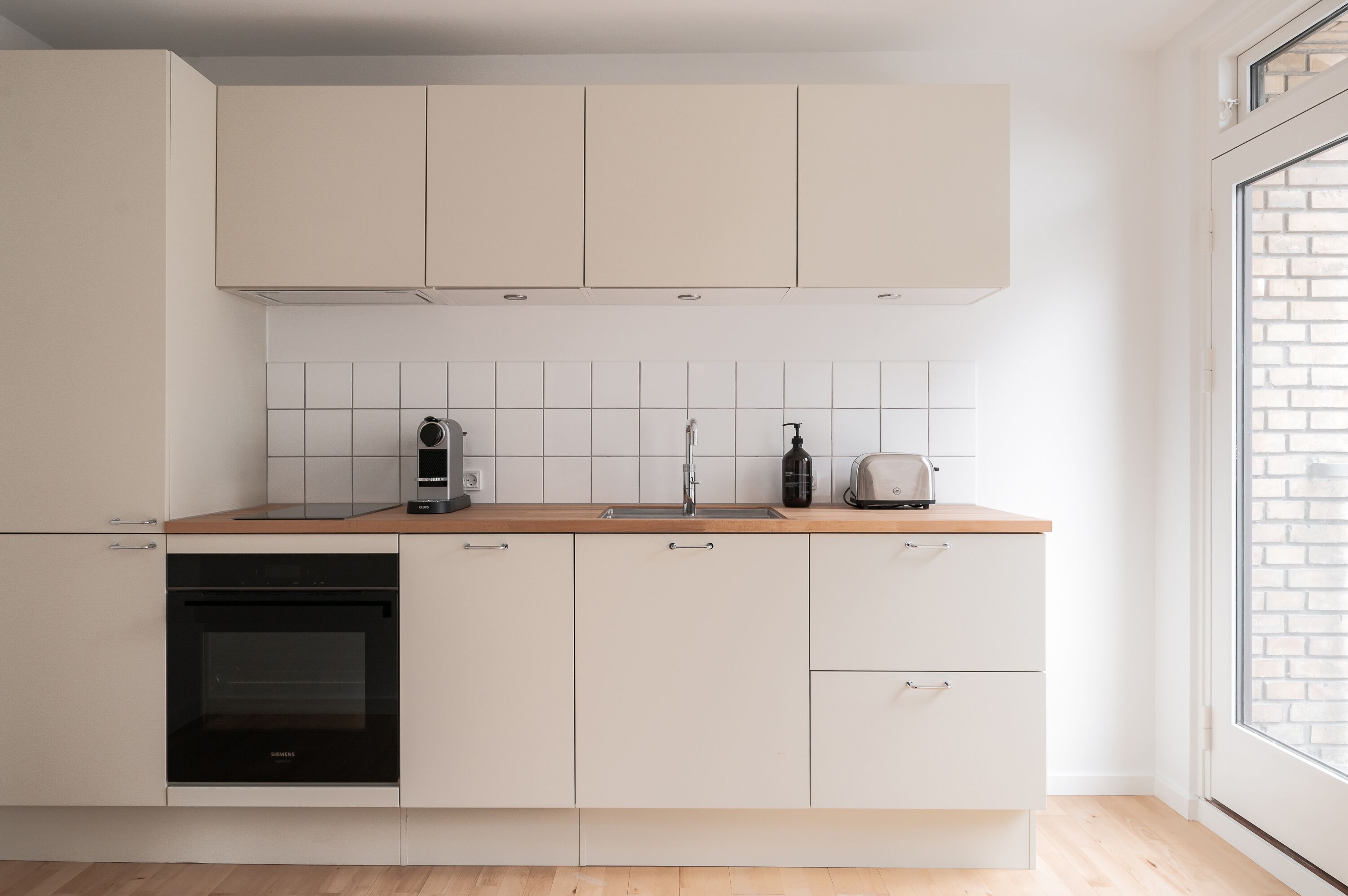 Eine moderne Küche mit weißen Schränken, einem schwarzen Backofen, einer Holz Arbeitsplatte und einem Fenster mit Blick auf eine Backsteinmauer.