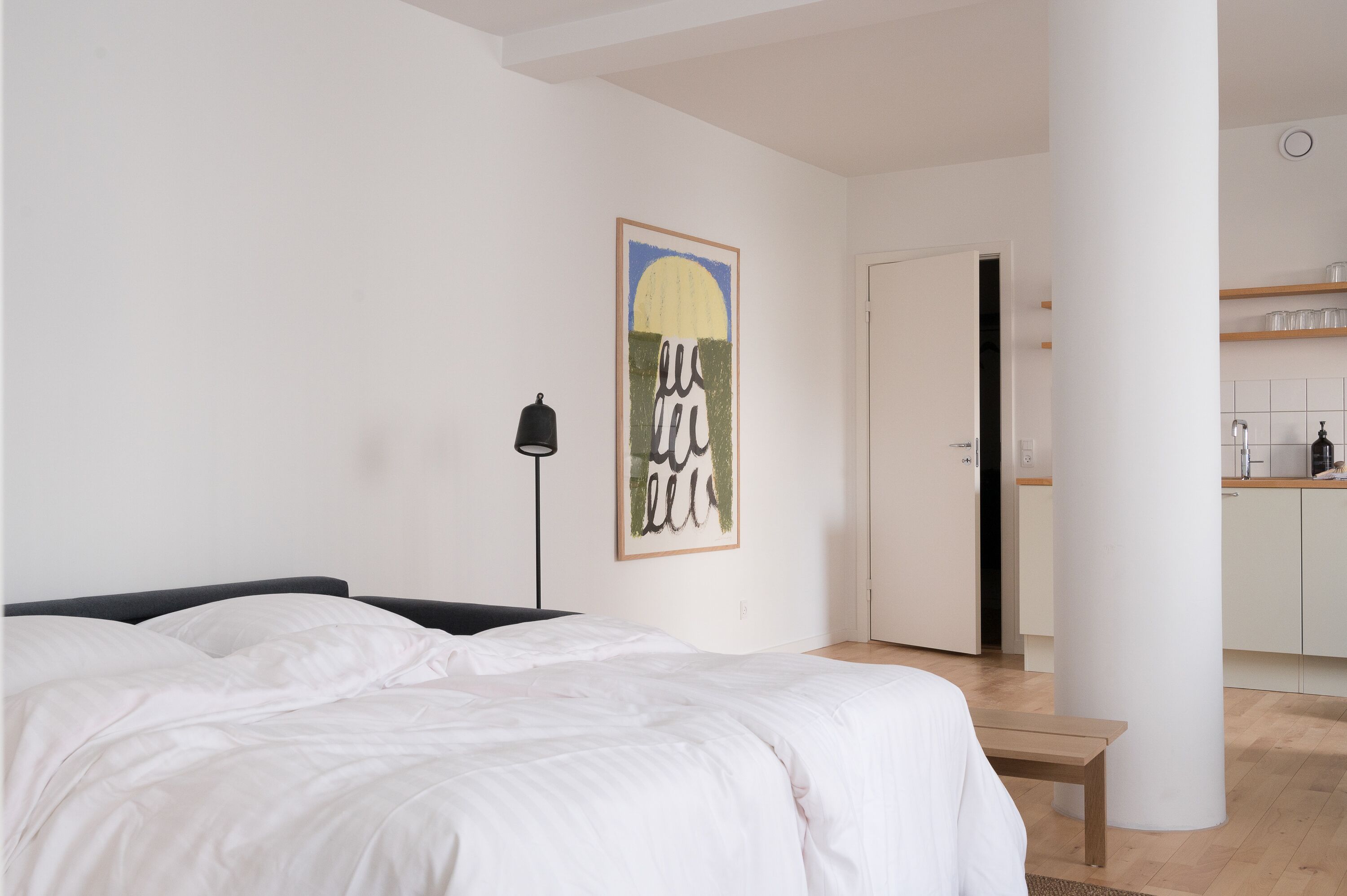 A bedroom with a bed, a bedside lamp, a wall art piece, and a view into a kitchen area.