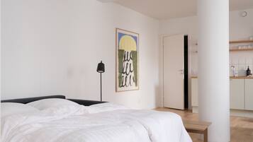 A bedroom with a bed, a bedside lamp, a wall art piece, and a view into a kitchen area.
