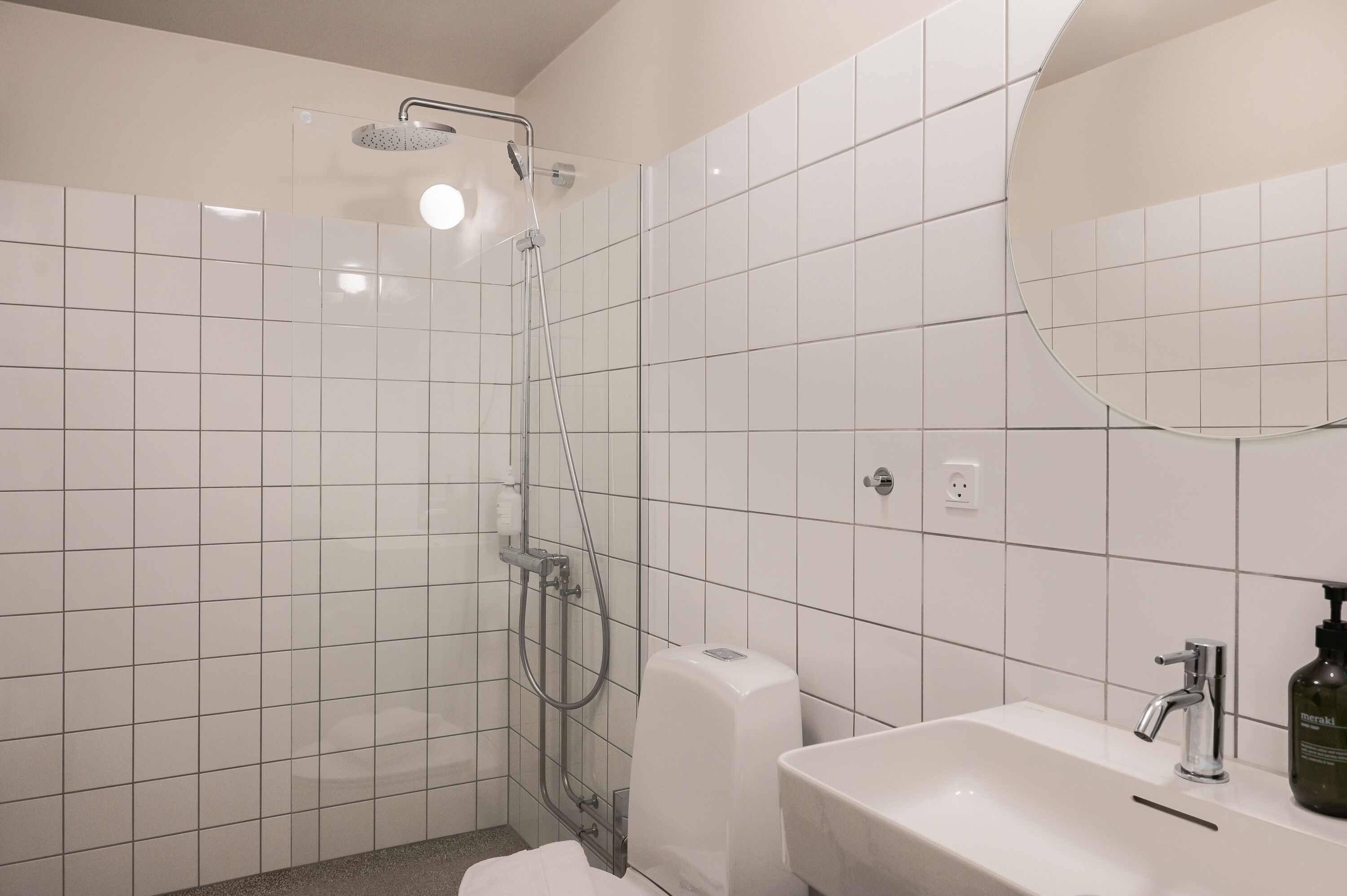 Un baño moderno con azulejos blancos, una ducha con cabezal de ducha manual, un inodoro y un lavamanos con un espejo arriba.