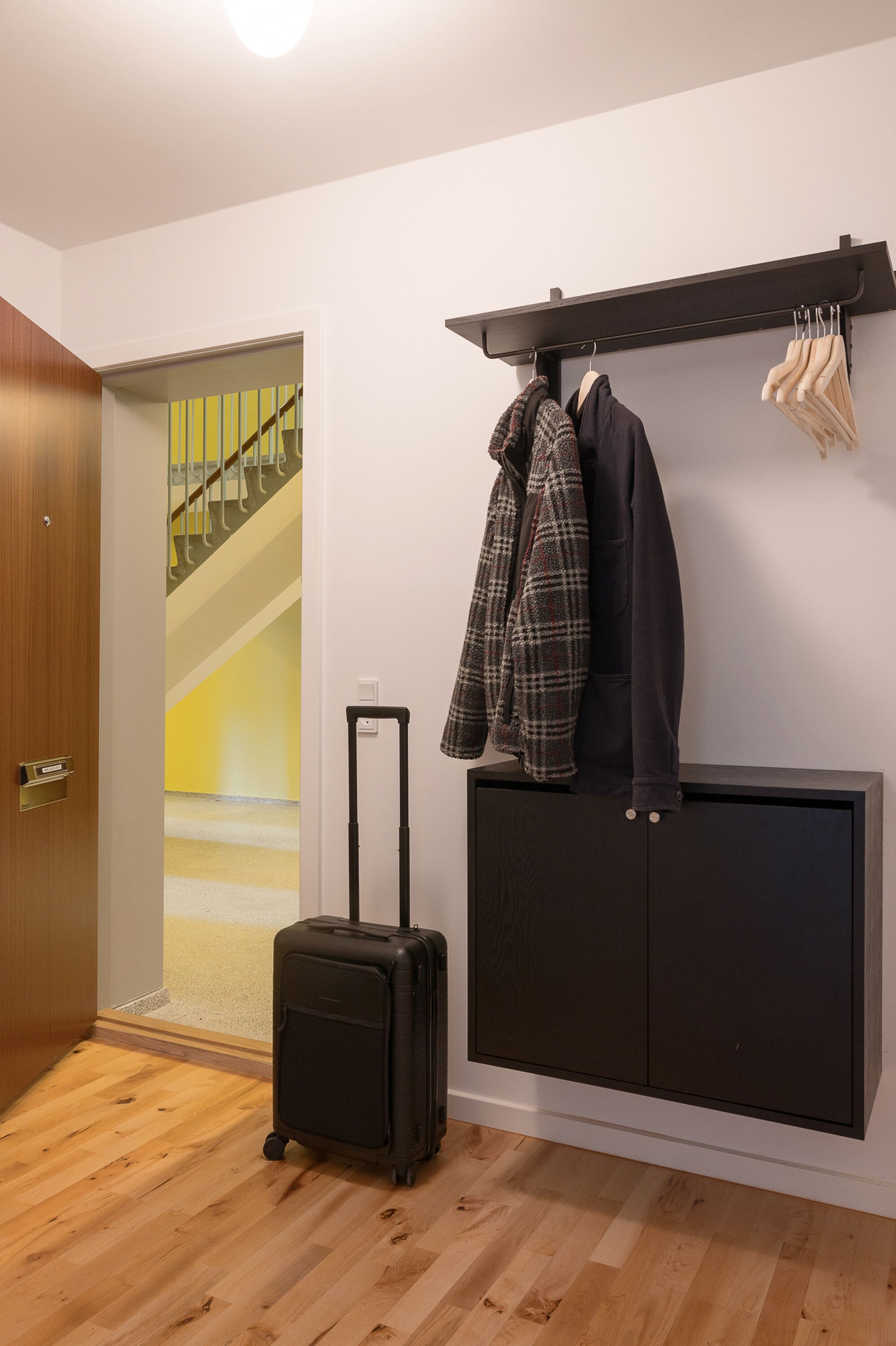 Ein Zimmer mit Holzboden, einem schwarzen Koffer, einem Kleiderständer mit Jacken und einer Tür zu einer Treppe.