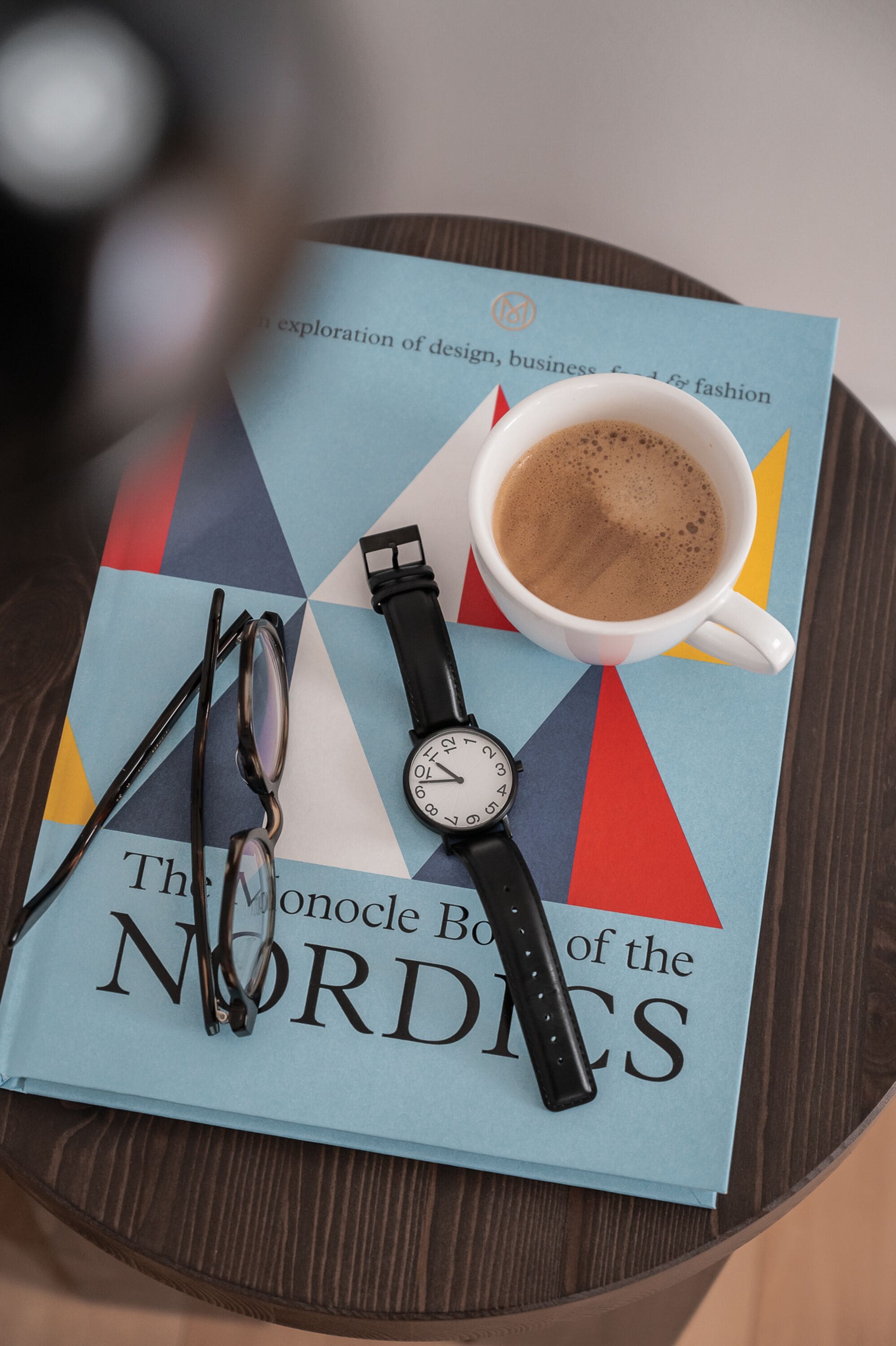 A book titled "The Monocle Book of the Nordics" on a table with a cup of coffee, glasses, and a watch.