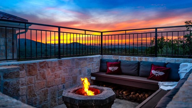 A stone fire pit with a fire, a stone bench, and a cushioned seating area with pillows, all enclosed by a metal railing with a sunset view. - Villa Olivella with Heated Pool (Vodice)