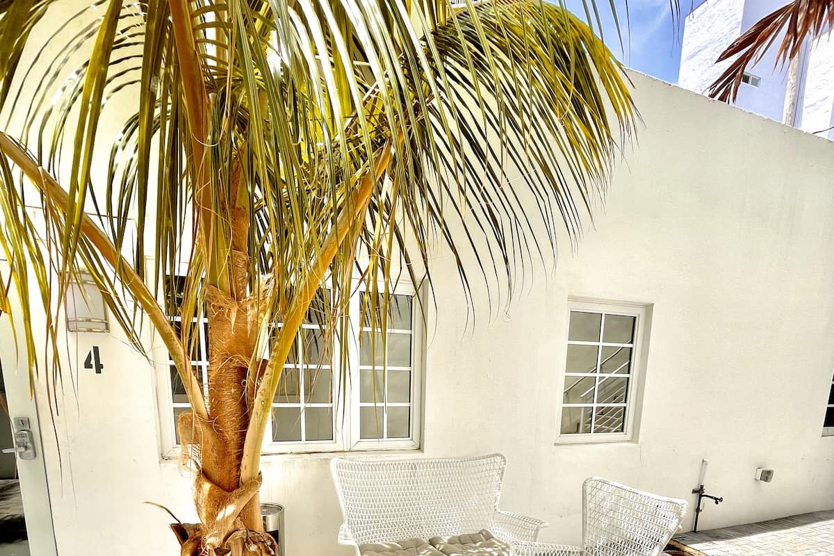 Un edificio blanco con una palmera, dos sillas blancas y una mesa redonda pequeña.
