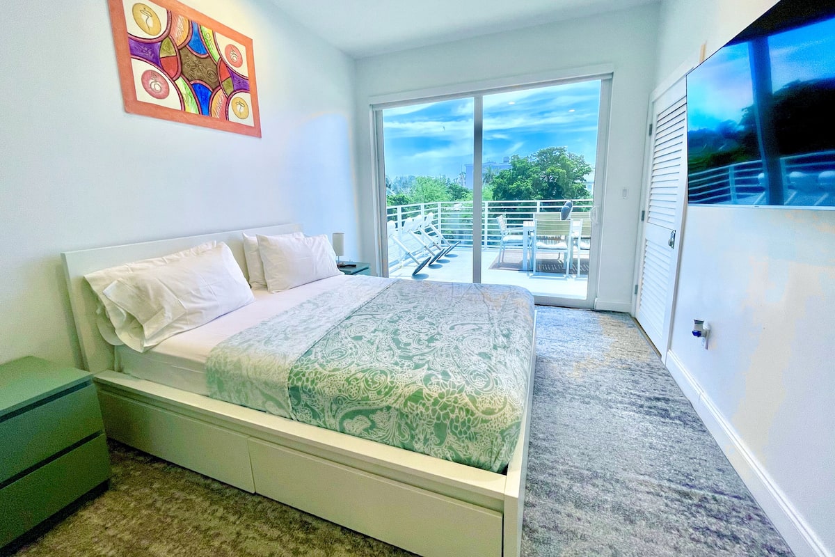 Un dormitorio con cama, mesita de noche, cómoda, ventana grande y balcón con mobiliario exterior.