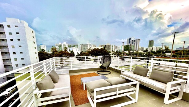 Une terrasse sur le toit, meublée de mobilier blanc, notamment des canapés et une table, d’où l’on peut admirer un panorama sur la ville, avec ses bâtiments modernes.