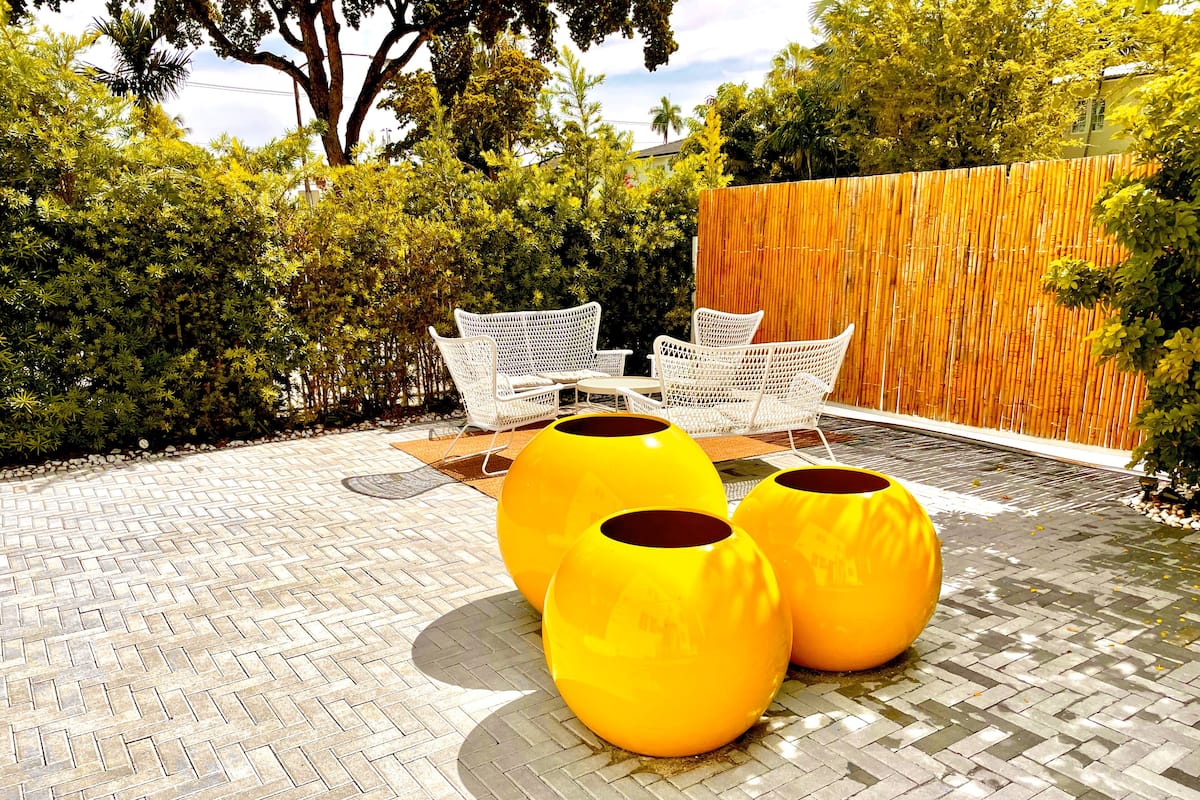 Un patio con suelo de ladrillo estampado, mobiliario de mimbre blanco y tres macetas esféricas amarillas.