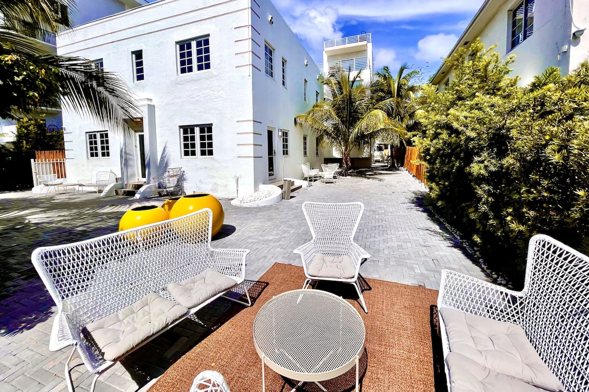 Un patio con mobiliario de mimbre blanco, una maceta amarilla y un edificio blanco con varias ventanas.