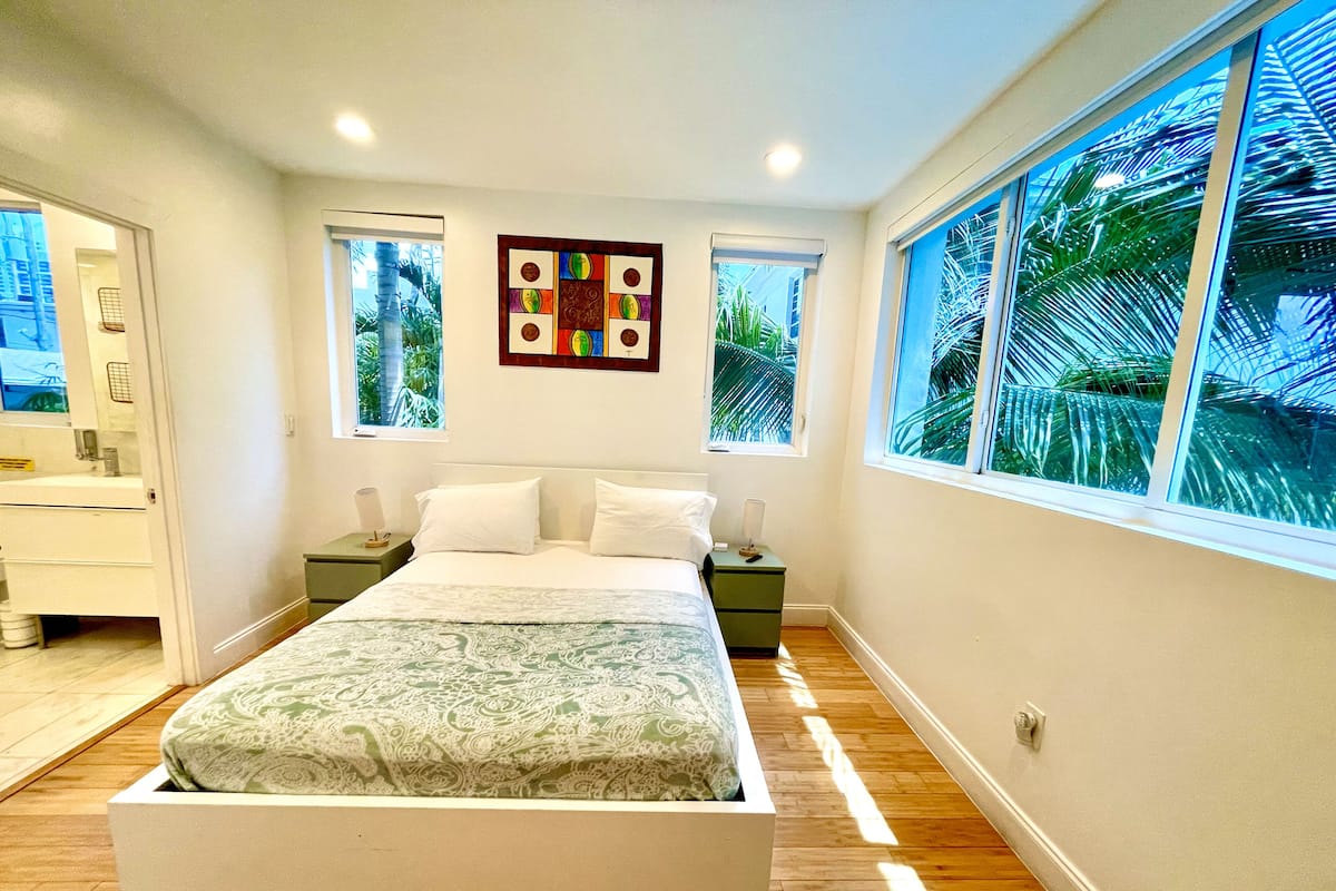 Un dormitorio moderno con una cama grande, dos mesitas de noche, una mesilla auxiliar, una obra de arte en la pared y una ventana con vistas a palmeras.