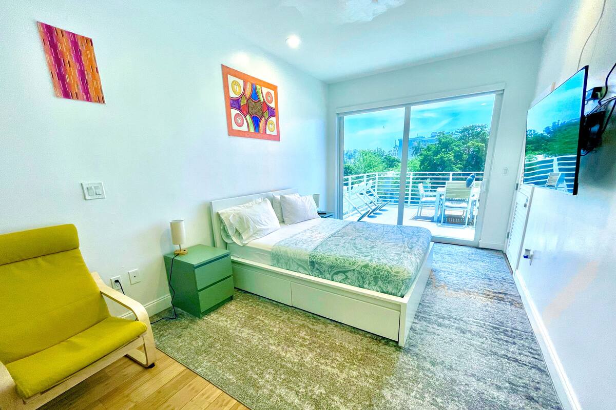 Un dormitorio moderno con una cama, una mesita de noche verde, un sillón amarillo y un balcón con vistas.