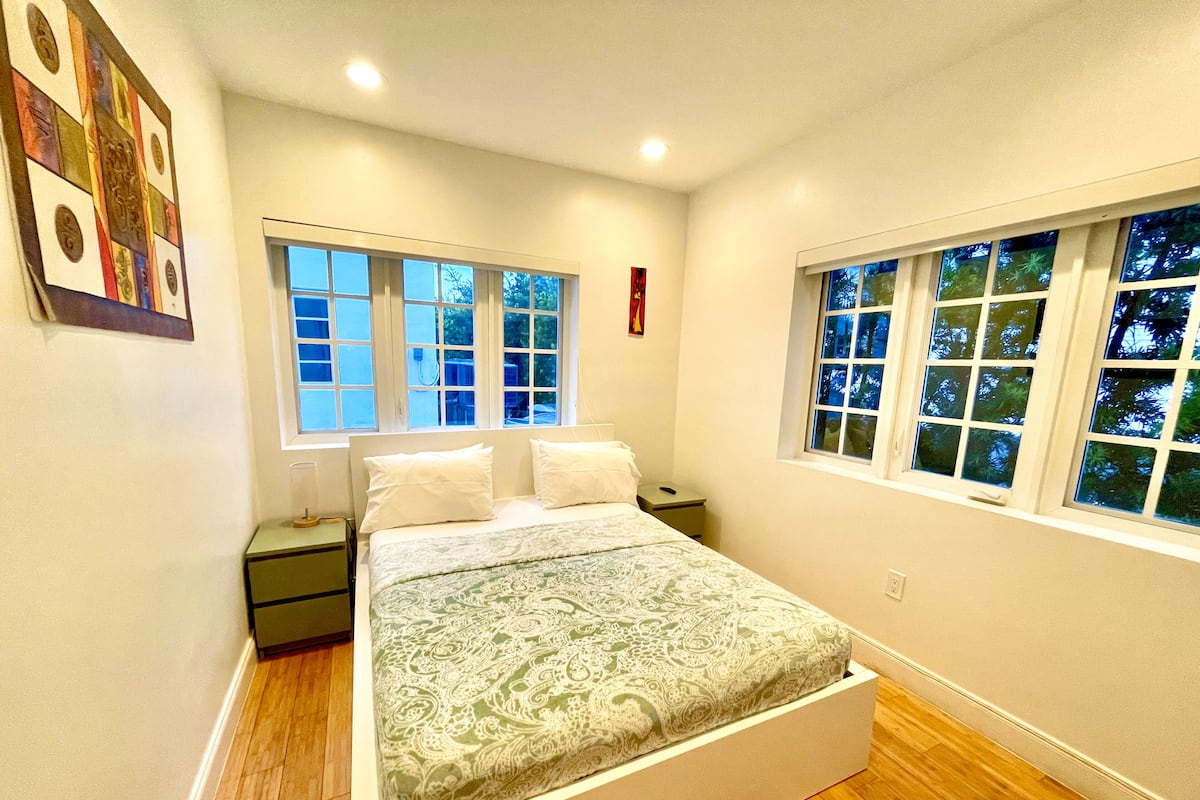 Un dormitorio con una cama, dos mesitas de noche y dos ventanas.