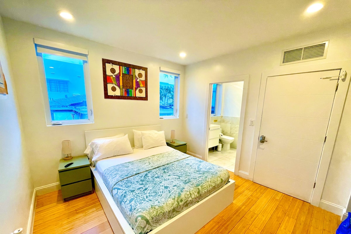 Un dormitorio con cama, mesitas de noche, una ventana, una puerta y un baño visible a través de una puerta entreabierta.