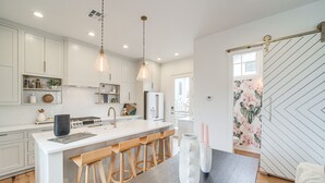 Une cuisine moderne avec des meubles blancs, des tabourets en bois, un îlot central et des luminaires suspendus.