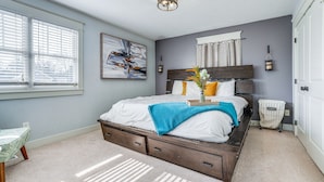 A bedroom with a large bed, a nightstand, a wall-mounted lamp, and a window with blinds. - Charming Kalamazoo Home w/ Yard: 1 Mi to Downtown! (Kalamazoo)