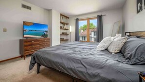 A bedroom with a large bed, a TV on a wooden dresser, a window with curtains, and a view of a house outside. - Family-Friendly 2BR Condo w/ Private Garage - Minutes from Zoo & Downtown (Fort Wayne)