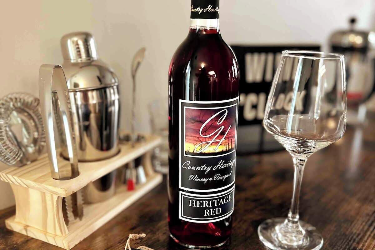A bottle of Heritage Red wine with a label, a wine glass, and a cocktail shaker on a wooden surface.