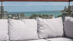 A balcony with a metal railing, a cushioned seating area with pillows, and a view of the sea and buildings in the background. - The Mordon (Newquay)