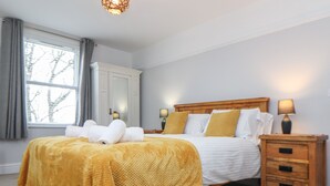 A bedroom with a large bed, a wooden nightstand, a bedside table, and a window with curtains. - The Mordon (Newquay)