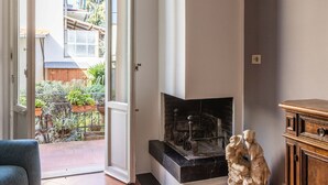 Ein Zimmer mit Kamin, einem Holzkasten und Blick auf einen Garten durch eine offene Tür.