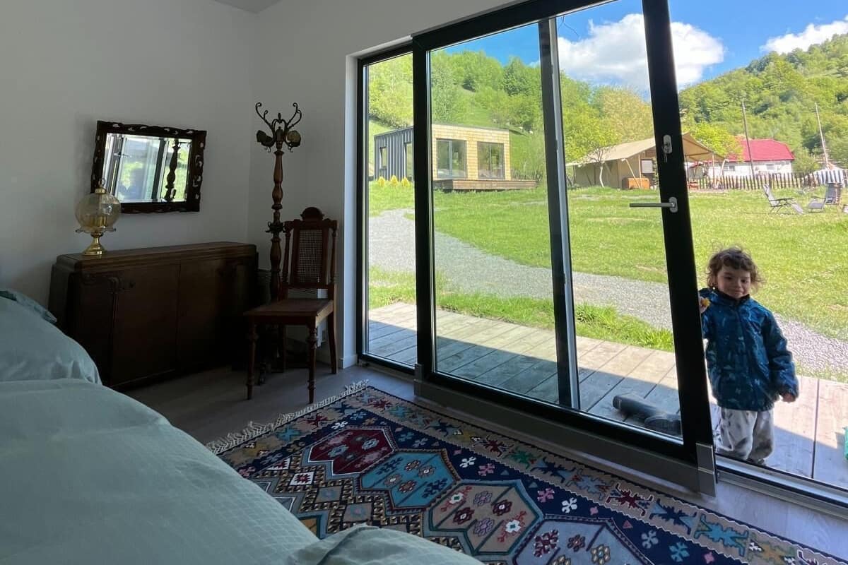 A bedroom with a large window, a bed, a chair, a wooden dresser, and a patterned rug.