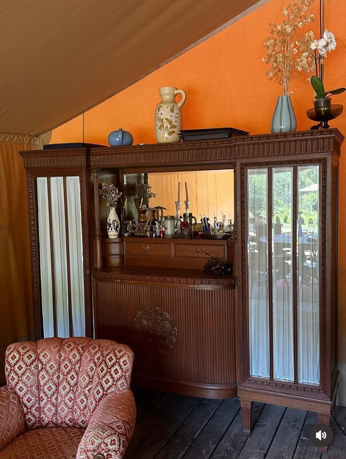 Un aparador tradicional de madera con puertas de cristal, un sillón estampado y una mesita con un jarrón de flores.
