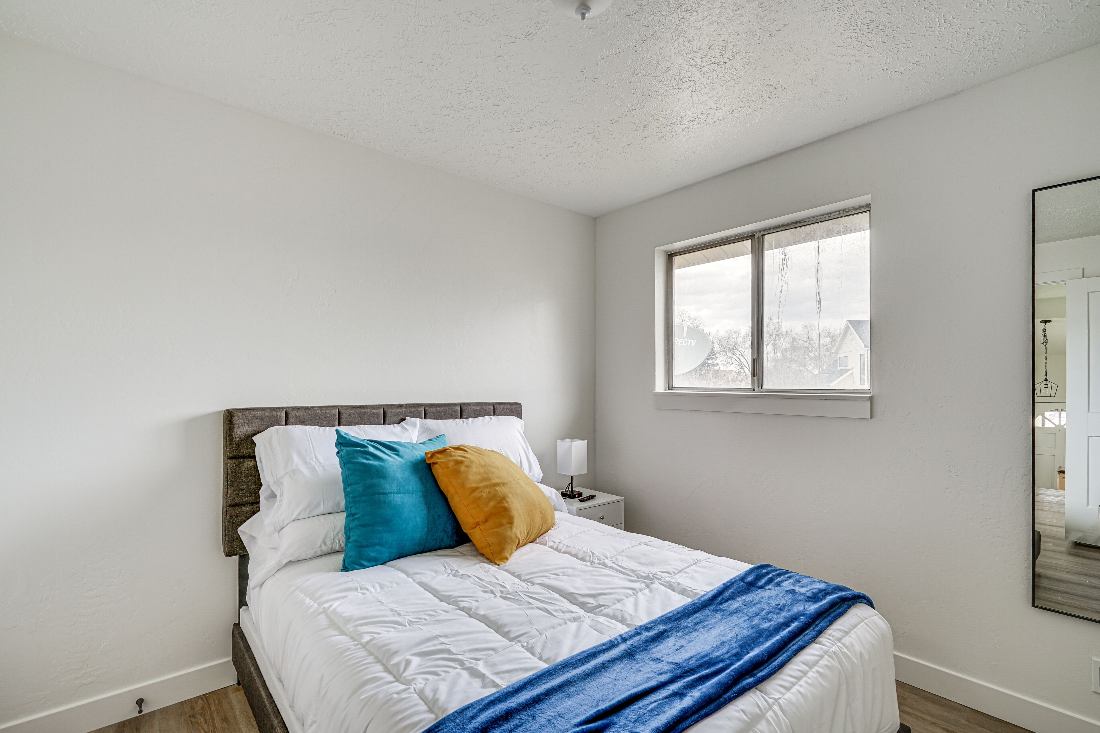 A bedroom with a bed, white walls, a window, and a nightstand with a lamp.