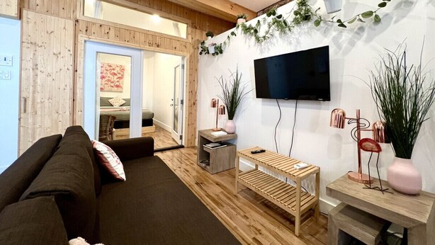 A modern living room with a wooden floor, a large flat-screen TV mounted on the wall, a sofa, a coffee table, and a small side table with a lamp. - The Flamingo ~ Cozy appartement in Saint-Roch - Downtown Quebec (Québec)