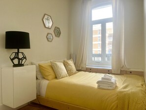Une chambre à coucher avec un lit, une table de chevet, une lampe et une fenêtre avec des rideaux.