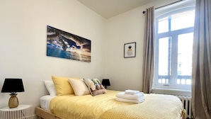 A bedroom with a wooden bed, white bedding, a tropical-themed painting, a framed pineapple picture, and a window with curtains. - L'Ananas ~ A Charming Home in the Heart of Downtown (Québec)