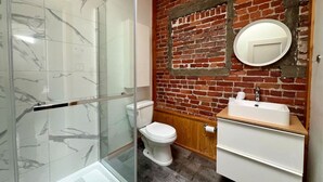 A bathroom with a glass shower enclosure, a white toilet, a wooden vanity with a sink, and a brick wall. - The Cactus ~ Superb accomodation in Saint-Roch, Downtown Quebec (Québec)