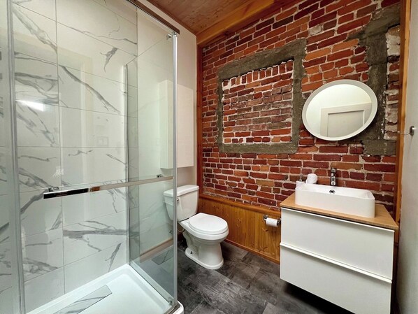 Une salle de bain avec une douche à parois de verre, des toilettes blanches, un meuble-lavabo en bois et un mur en briques.