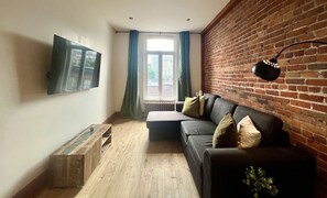 A living room with a brick wall, a black leather sofa, a wooden coffee table, and a flat-screen TV mounted on the wall. - Le Cactus ~ A Stunning Home in the St-Roch Neighborhood (Québec)