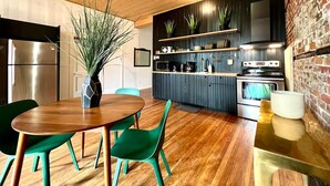 A modern kitchen with wooden flooring, a dining area with a wooden table and green chairs, and a brick wall.