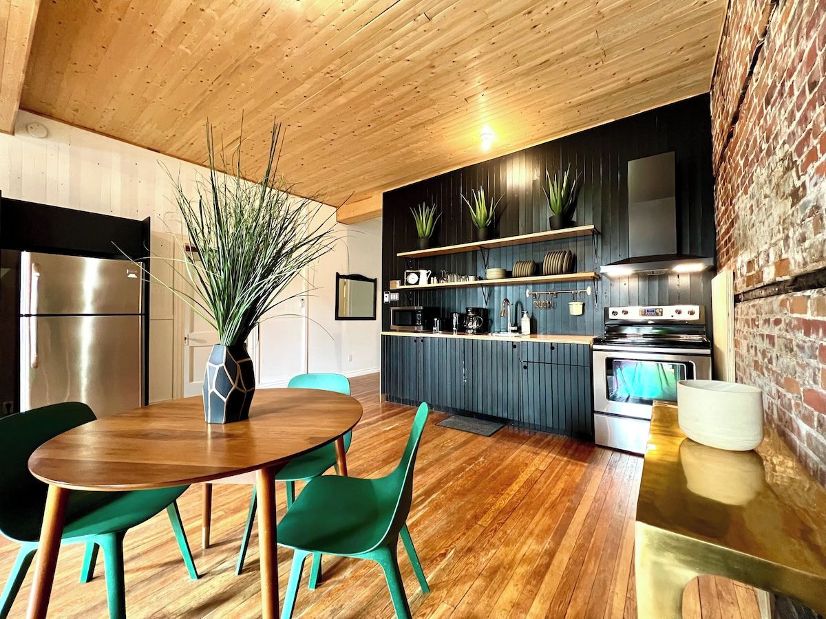 Una cocina moderna con piso de madera, una zona de comedor con mesa de madera y sillas verdes, y una pared de ladrillo.