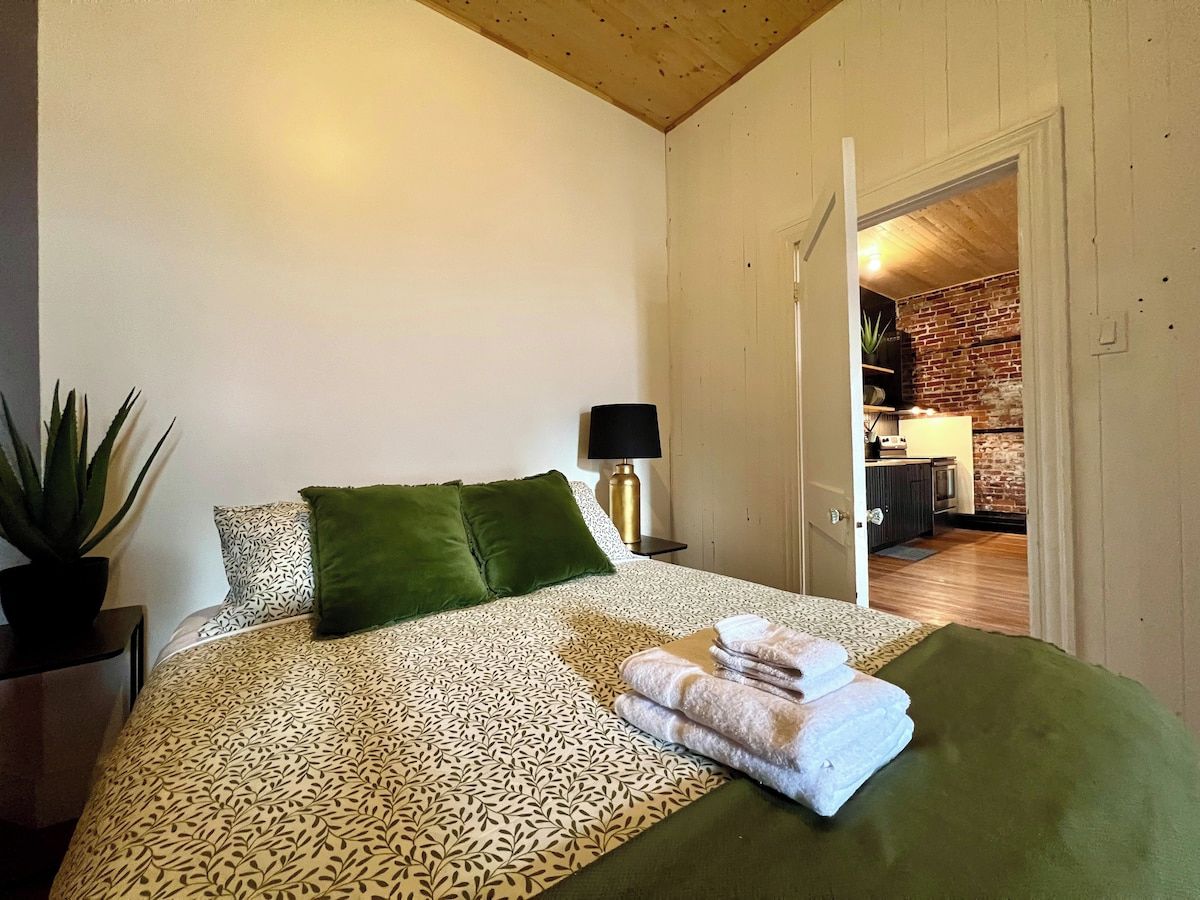 Un dormitorio con una colcha estampada, almohadas verdes, una lámpara de noche y una toalla doblada cuidadosamente sobre la cama.