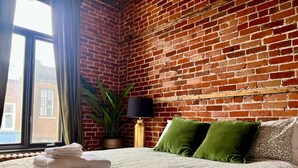 A hotel room with a brick wall, a bed with a patterned bedspread, green pillows, a lamp, and a plant.