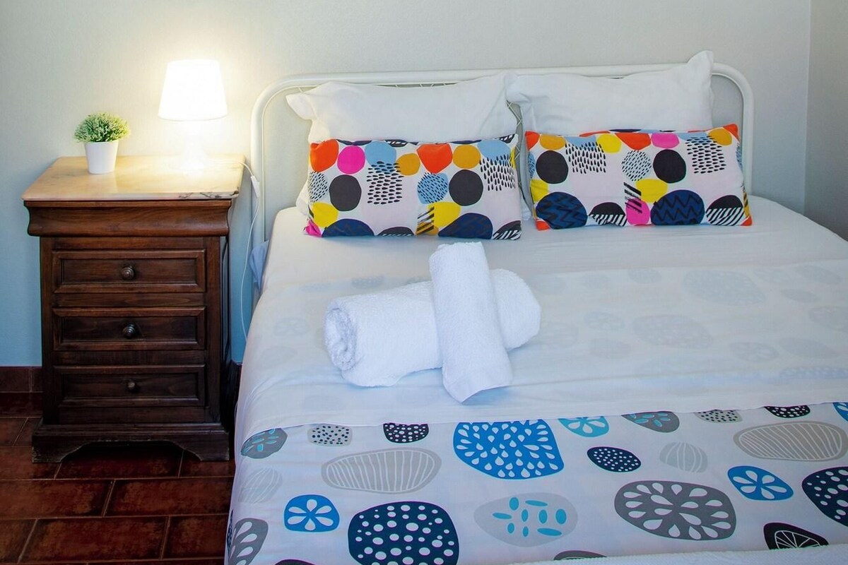 Een slaapkamer met een bed, kussens, een nachtkastje, een lamp en een spiegel aan de muur.