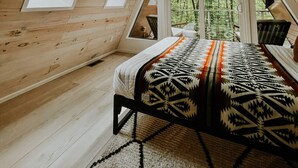 A cozy attic bedroom with wooden walls, a bed with a patterned blanket, and large windows offering a view of the forest. - Fox Ridge-Black Alder Lodging (Logan)